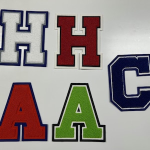 以下が含まれることがあります： さまざまな色の大学文字パッチのコレクション。文字には、2つの「A」、2つの「H」、および1つの「C」が含まれます。文字は赤、緑、白、ネイビーブルーで、コントラストのあるボーダーが付いています。パッチは、おそらくアパレルをカスタマイズするためのものです。