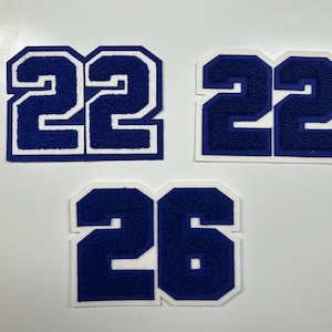 以下が含まれることがあります： 青と白の大学番号パッチが3つ。2つのパッチには数字の22が表示され、3つ目のパッチには数字の26が表示されています。数字は、白い縁取りのある太字のブロック体フォントです。