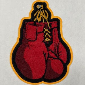 May include: A red boxing glove patch with black outlines and a gold border. The gloves are laced together with yellow laces. The patch is a sports-themed item.