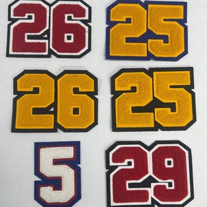 May include: A collection of six embroidered patches featuring two-digit numbers. The patches include the numbers 26 in maroon, 25 in gold, 26 in gold, 25 in gold, 5 in white, and 29 in maroon. Each patch has a thick, contrasting border.