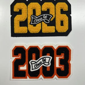 以下が含まれることがあります： 2026年と2003年の刺繍パッチが2つ。2026年のパッチは、黒い背景に金色の数字があり、「Class Of」がバナーに表示されています。2003年のパッチは、オレンジと白の背景に黒い数字があり、「Class Of」がバナーに表示されています。