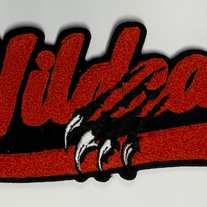 Pode incluir: Etiqueta de feltro vermelho e preto com a palavra "Wildcats" em uma fonte estilizada. Uma pegada de pata preta com garras brancas está centralizada abaixo da palavra.