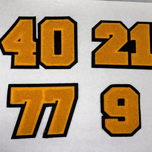 以下が含まれることがあります： 黒い輪郭と金色のフェルトでできた4つのフェルトパッチ。数字の40、21、77、9が見えます。