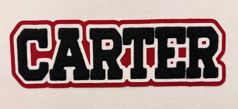 May include: A fabric patch featuring the name CARTER in bold black block letters. The lettering is outlined in a thick white border, which is then surrounded by a vibrant red background layer that follows the contour of the text.