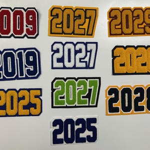 以下が含まれることがあります： 2009年、2019年、2025年、2026年、2027年、2028年、2029年と書かれた、カラフルなフェルトパッチのコレクション。パッチは、赤、青、金、緑など、さまざまな色の組み合わせの、大学の文字スタイルです。