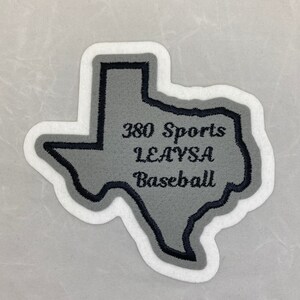 Peut inclure: Un écusson en feutre gris en forme de l'état du Texas avec des contours brodés noirs. L'écusson porte le texte "380 Sports Leavsa Baseball" brodé en noir.