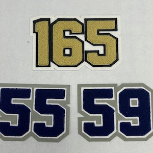 May include: Four felt patches with numbers on them. The top patch is gold with the numbers 165. The bottom two patches are gray with navy blue numbers. The numbers are 55 and 59.
