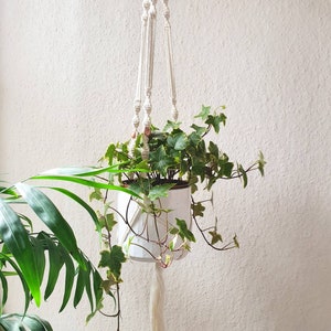 Op de afbeelding: Een witte hangende plantenpot met een macramé-ontwerp en koperen kralen. De pot is gevuld met groene klimop planten.