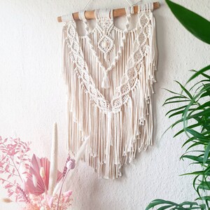 May include: A white macrame wall hanging with a wooden dowel at the top. The hanging features a geometric pattern with fringe details.