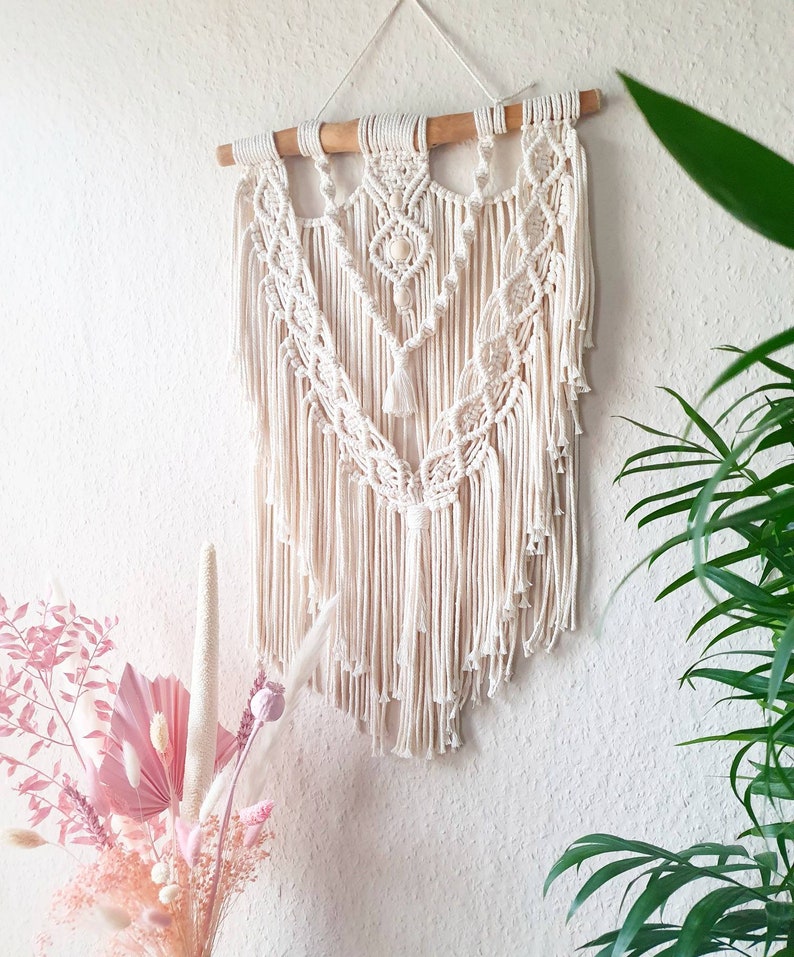 May include: A white macrame wall hanging with a wooden dowel at the top. The hanging features a geometric pattern with fringe details.