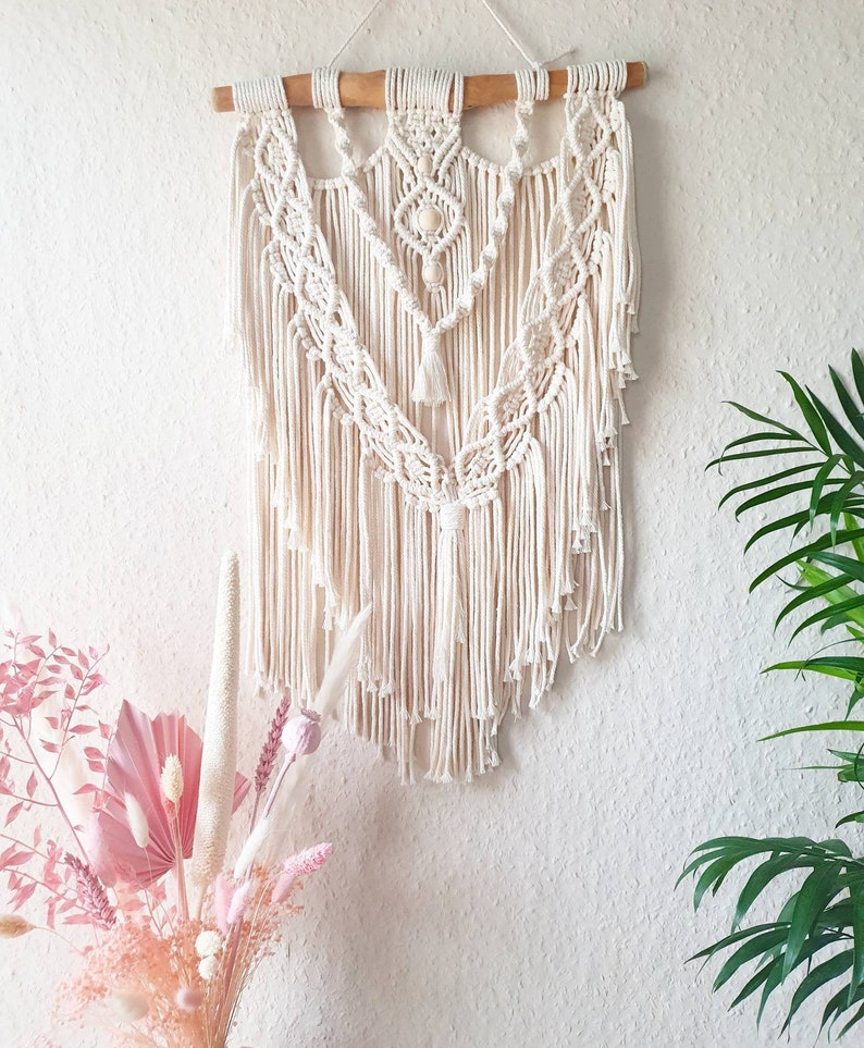 May include: A white macrame wall hanging with a wooden dowel at the top. The macrame features a geometric pattern with fringe and beads.