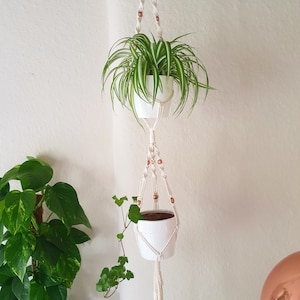 Op de afbeelding: Een witte macrame plantenhanger met koperen kralen houdt twee witte potten met groene planten vast. De hanger is opgehangen aan een haak aan het plafond.
