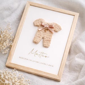 Può includere: Opera d'arte incorniciata con un body per neonati all'uncinetto beige, fiocco rosa e campanella. Il nome "Matteo" è scritto in oro, con dettagli di nascita: 01.02.2026, 01.24, 2.635g e 49cm. La cornice è in legno chiaro. Il testo "lively swan" è in basso.