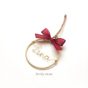 May include: Gold wire ornament with the name "Lena" written in cursive. The ornament is decorated with a burgundy ribbon bow and a twine hanger.
