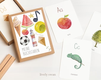 Könnte beinhalten: Ein Set bunter Alphabet-Karteikarten mit Aquarell-Illustrationen verschiedener Gegenstände. Die Karten zeigen die Buchstaben A bis D und die deutschen Wörter für Apfel, Baum und Chamäleon. Die Schachtel zeigt verschiedene Illustrationen, darunter einen Fußball, ein Eis am Stiel, eine Wassermelone, einen Leuchtturm und einen Drachen. Der Text auf der Schachtel lautet "Meine Buchstaben".