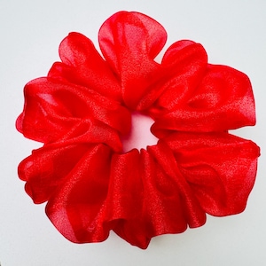 May include: A vibrant red scrunchie made of sheer fabric. The scrunchie is gathered and ruffled, creating a voluminous, flower-like shape. The bright red color is consistent throughout the fabric, and the scrunchie is set against a white background.