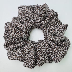 Peut inclure: Grosse chouchou avec un imprimé léopard. Le tissu présente un motif de taches marron, beige et crème. La chouchou est circulaire et a un aspect froncé et froissé, adapté à diverses coiffures.