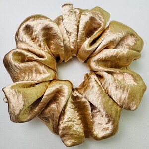 May include: A gold-colored scrunchie made of a soft, velvety fabric. The scrunchie is circular, with multiple folds of fabric creating a voluminous, textured appearance. The color is a warm, metallic gold, reflecting light.