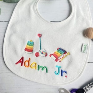 Puede incluir: Babero blanco para bebé con diseños de juguetes bordados coloridos, incluyendo una torre apilable, un juguete para tirar y un xilófono. El nombre "Adam Jr." está bordado en colores arcoíris. Se ve una sonaja de madera.