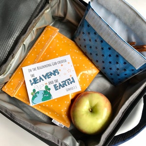 May include: A blue lunch bag with a zipper closure. Inside the bag is a yellow lunch bag with white polka dots, a blue lunch bag with white stars, a red apple, and a card with the text "In the beginning God created the heavens and the earth Genesis 1:1".