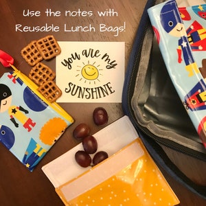 Pode incluir: Uma bolsa de almo&ccedil;o reutiliz&aacute;vel azul e amarela com um design de super-her&oacute;i. A bolsa est&aacute; aberta e mostra uma nota que diz "You are my sunshine" com um rosto sorridente. Tamb&eacute;m h&aacute; alguns pretzels e uvas dentro da bolsa.