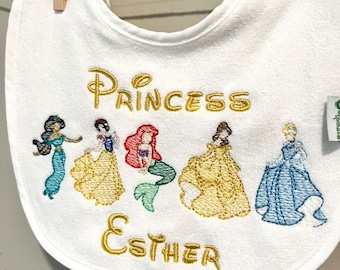 May include: White baby bib embroidered with the word "Princess" and the name "Esther" in gold thread. The bib features embroidered images of Disney princesses in various colors, including blue, yellow, and red. The bib is hanging from a clothes pin.