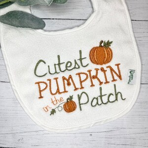 May include: White baby bib embroidered with the phrase "Cutest PUMPKIN in the Patch" in green and orange thread. The bib features two embroidered pumpkins, one large and one small, with green leaves.