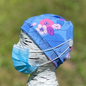 May include: A blue floral patterned scrub cap with pink and white flowers and green leaves. The cap has a white elastic band and a button on the side to hold a blue face mask. The background is a blurred green.