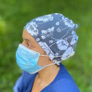 May include: A gray and white floral scrub cap with a white button on the side. The cap is being worn by a person wearing a blue surgical mask.