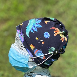 May include: Black scrub hat with a colorful dinosaur and space-themed print. The hat has a button on the side for attaching a face mask.