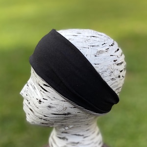 May include: A black headband is displayed on a white mannequin head. The headband is wide and appears to be made of a soft, stretchy material. The background is a blurred green, suggesting an outdoor setting.