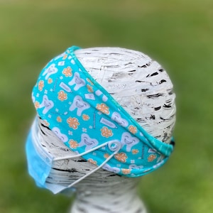May include: A teal headband with a pattern of smiling teeth, toothbrushes, and flowers. The headband is designed to hold a face mask in place with buttons. The mask is light blue.