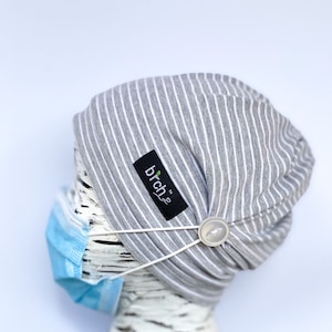 May include: Gray and white striped beanie with a white button and a black label that says "brch" with a green leaf logo. The beanie is on a white mannequin head with a blue face mask.