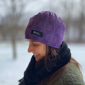 May include: A purple beanie with a geometric pattern in pink and light purple. The beanie has a small black tag with the word "birch" in white. The beanie is worn by a person in a winter setting.