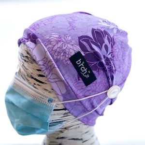 May include: Purple and white floral print scrub hat with a white button and elastic band. The hat has a black label with the text "brch2" on it.