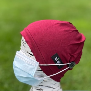 Puede incluir: Un gorro burdeos con un botón blanco y una correa elástica para sujetar una mascarilla facial. El gorro tiene un pequeño logotipo de hoja verde con la palabra "birch" en él.