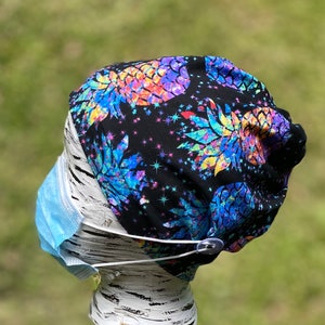 May include: A black scrub hat with a colorful pineapple print and a button for attaching a face mask. The hat has a white elastic band for a secure fit.