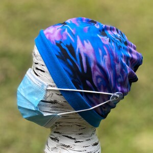 May include: A blue and purple tie-dye fabric face mask with a clear plastic button on the side. The mask is attached to a blue and purple tie-dye fabric headband.
