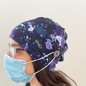 May include: A person wearing a blue and purple patterned scrub hat with a button on the side. The hat has a white elastic strap that goes around the back of the head and attaches to the button. The person is also wearing a blue surgical mask and glasses.