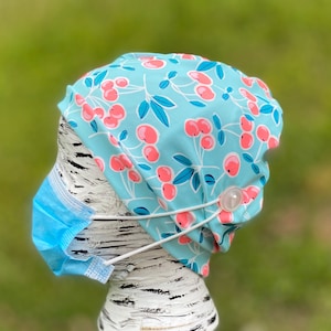 May include: A light blue scrub hat with a cherry print and a white button. The hat is on a white mannequin head.