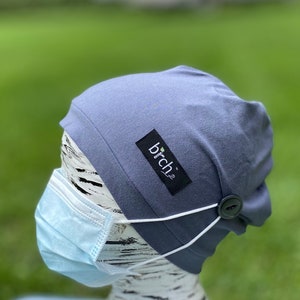 May include: A blue beanie with a button on the side and a white elastic strap. The beanie has a small black label with the word "birch" on it. The beanie is being worn on a mannequin with a blue surgical mask.