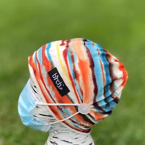 May include: A colorful scrub cap with a vibrant abstract pattern in orange, blue, and brown. The cap has a black label with the word "birch" on it. A light blue face mask is attached to the cap with a button.