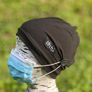 May include: A black beanie with a small label that says "birch." The beanie has a button on the side to hold the ear loops of a light blue face mask. The mask is in place, and the background is blurred green.
