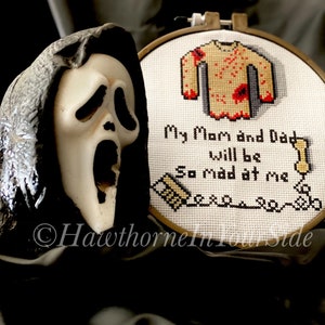 May include: A white embroidery hoop with a cross-stitch design of a bloody yellow sweater and the text "My Mom and Dad will be so mad at me". A white plastic Ghostface mask is in the foreground.