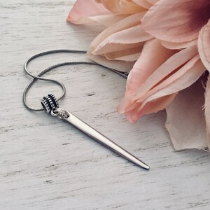 May include: A silver necklace with a long, thin, pointed pendant. The pendant is attached to the chain with a small, round bead. The necklace is lying on a white wooden surface.