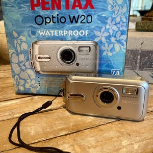 May include: A silver Pentax Optio w20 waterproof digital camera with a black wrist strap. The camera is sitting on a wooden surface. The box for the camera is in the background.