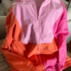 May include: A pink, orange, and hot pink color block windbreaker jacket with a zipper closure and ribbed cuffs.