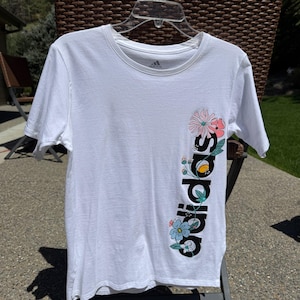 May include: White t-shirt with a black Adidas logo and floral design. The logo is vertical and the flowers are on the right side of the logo.