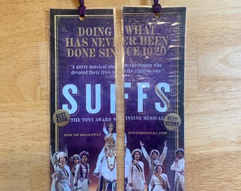 May include: Two rectangular paper bookmarks for the Suffs musical, featuring a purple background with white text reading Doing What Has Never Been Done Since 1920 and Suffs. Each bookmark has a purple tassel at the top and images of performers.