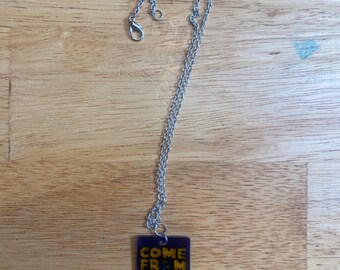 May include: A silver-toned metal chain necklace featuring a square, translucent purple pendant. The pendant displays the text "COME FROM AWAY" in bold, yellow, capitalized letters, with a small yellow airplane icon replacing the letter O.